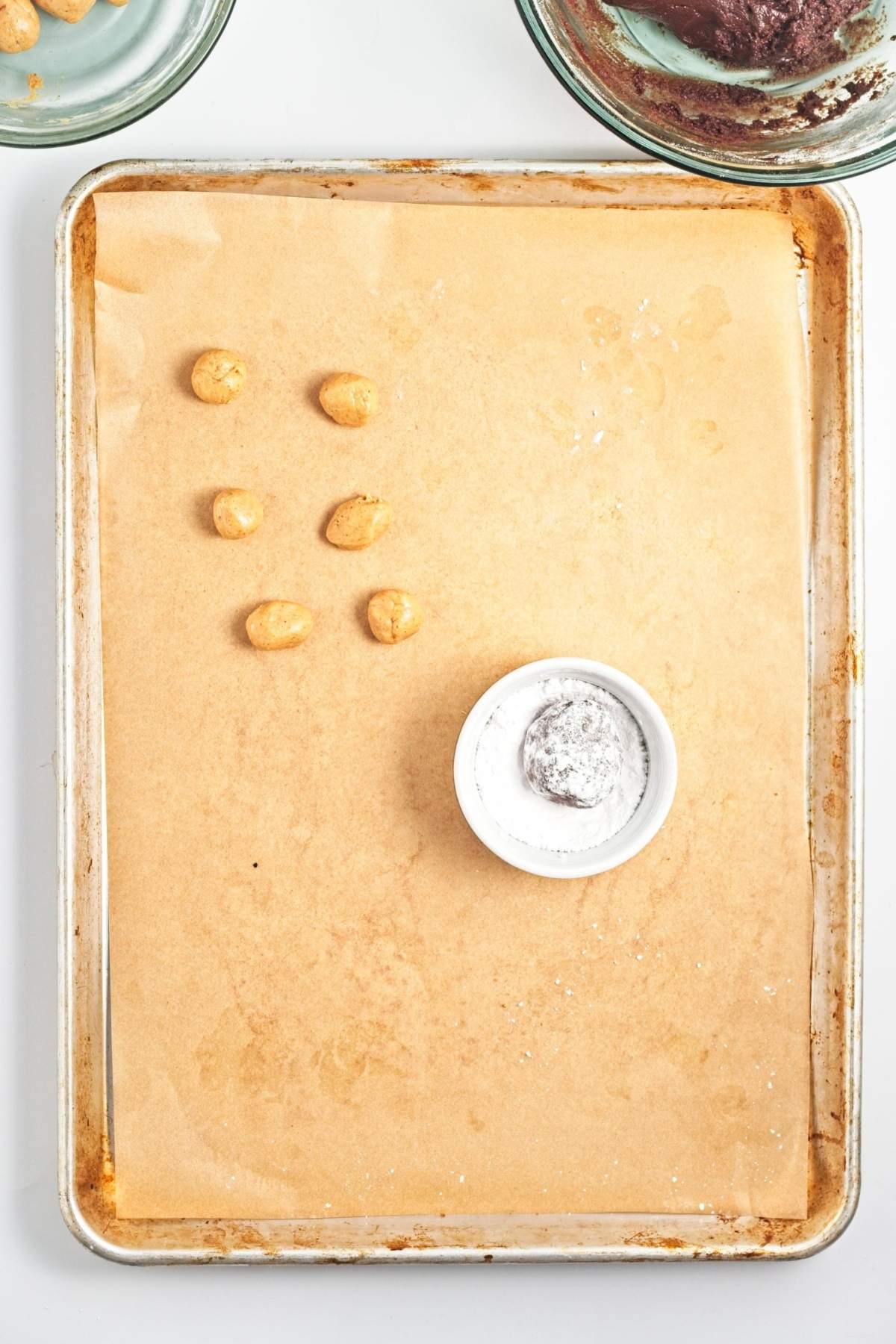 Peanut butter balls on a parchment-lined baking sheet, with one wrapped in chocolate dough and a small bowl of powdered sugar nearby.