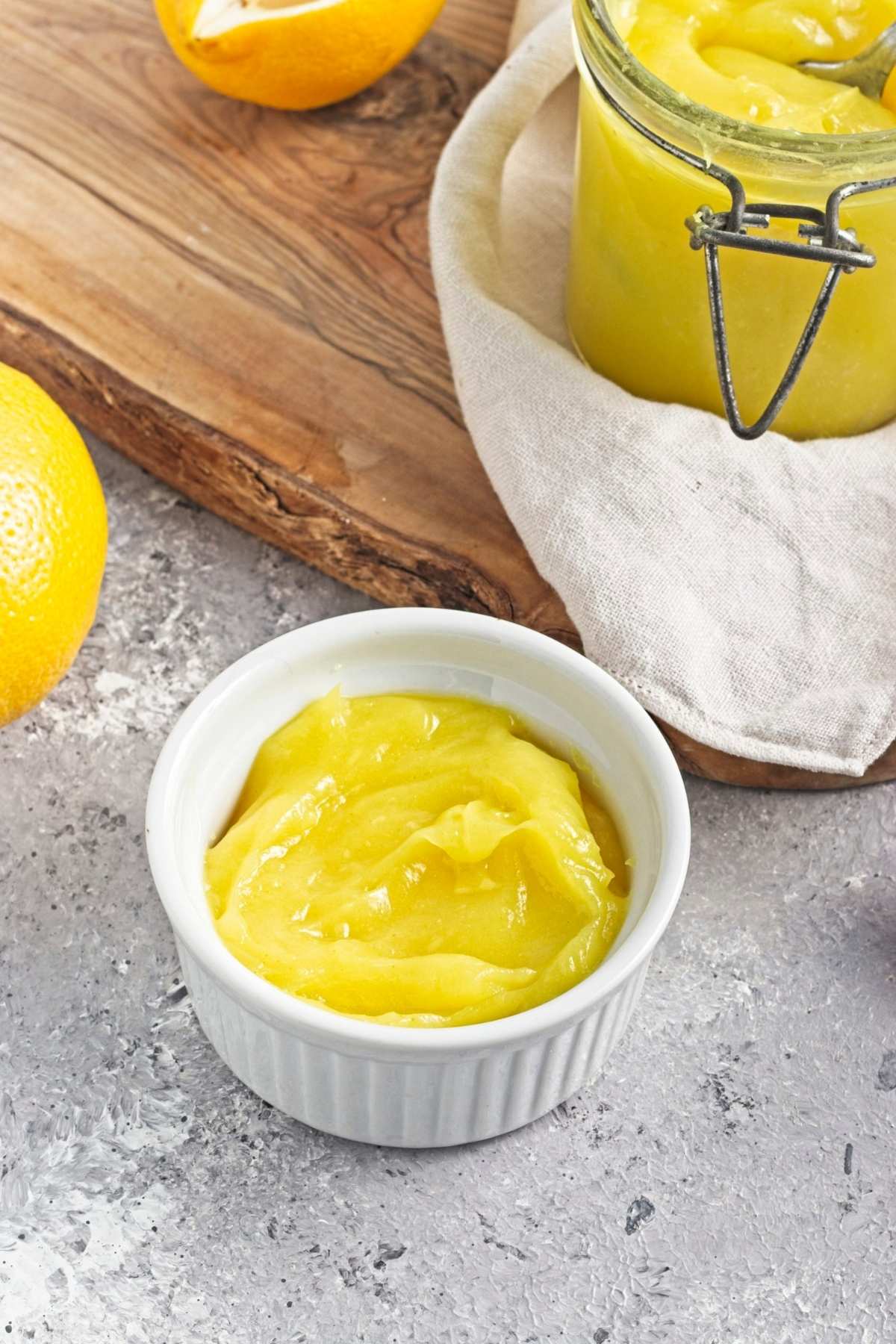 Small white ramekin of yellow curd beside a jar and halved lemons.