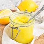 Open jar of yellow curd with spoon, with whole and halved lemons in the background.
