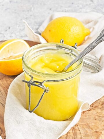 Open jar of yellow curd with spoon, with whole and halved lemons in the background.