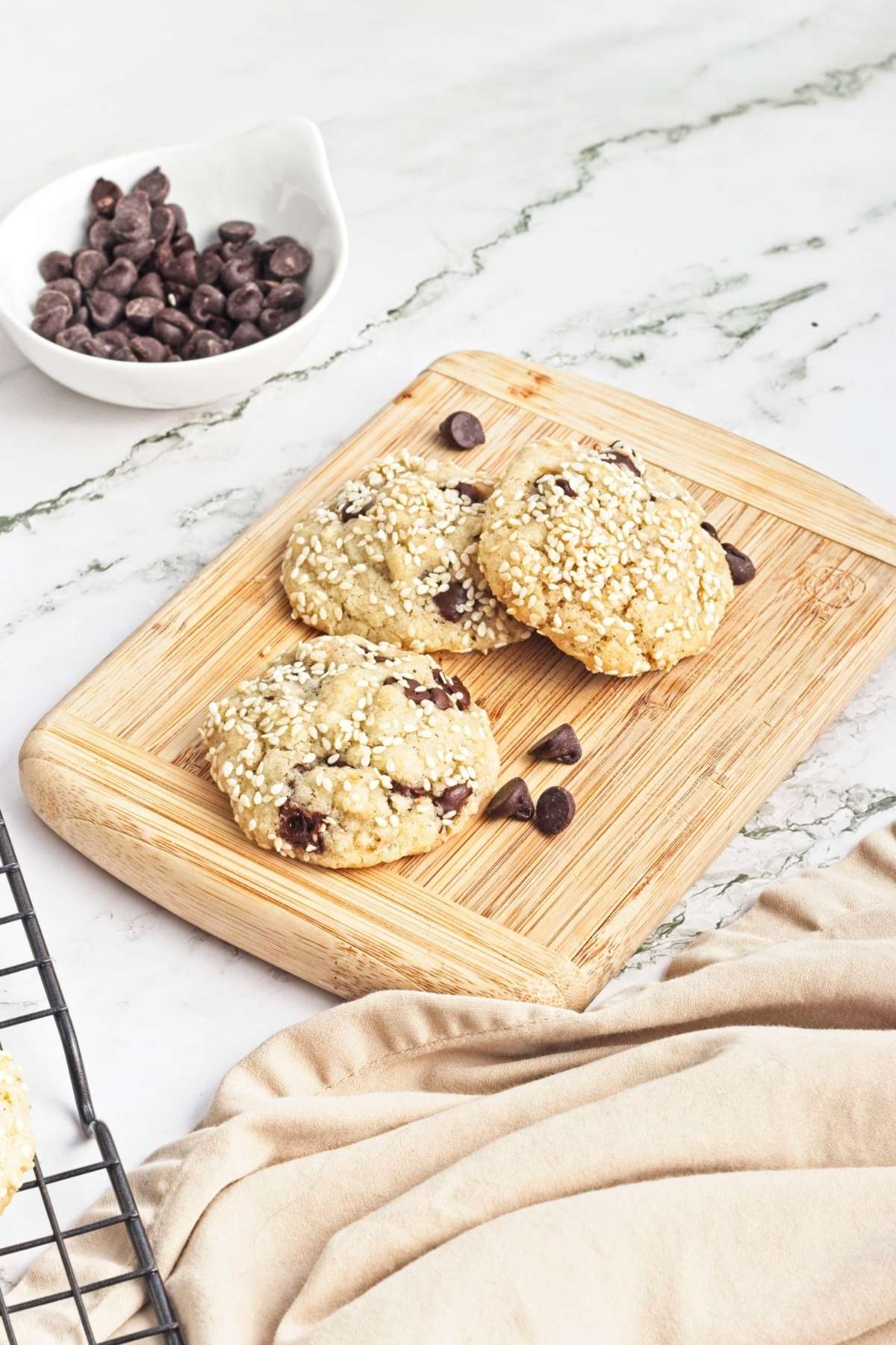 Three sesame-topped cookies with chocolate chips on a wooden board beside a bowl of chocolate chips.