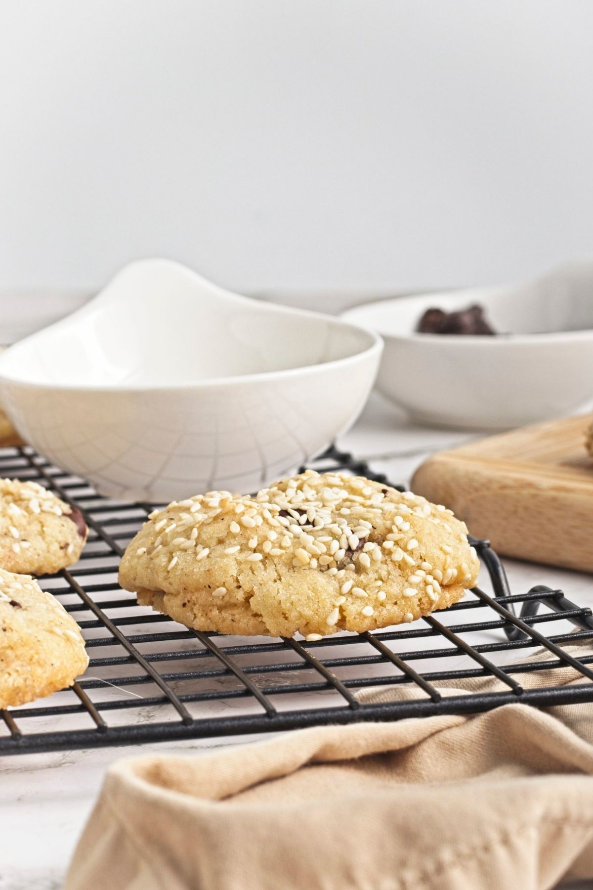 Close-up of a sesame-topped cookie cooling on a wire rack with bowls in the blurred background.