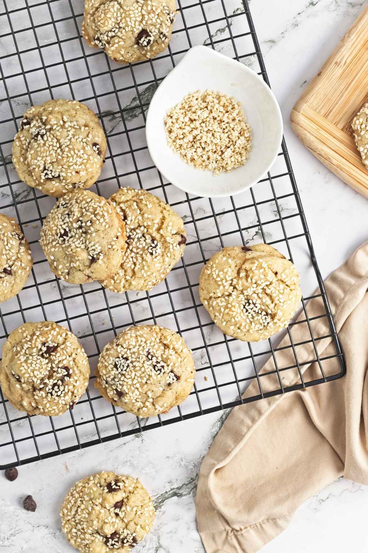 Freshly baked cookies topped with sesame seeds cooling on a wire rack beside a small bowl of sesame seeds.