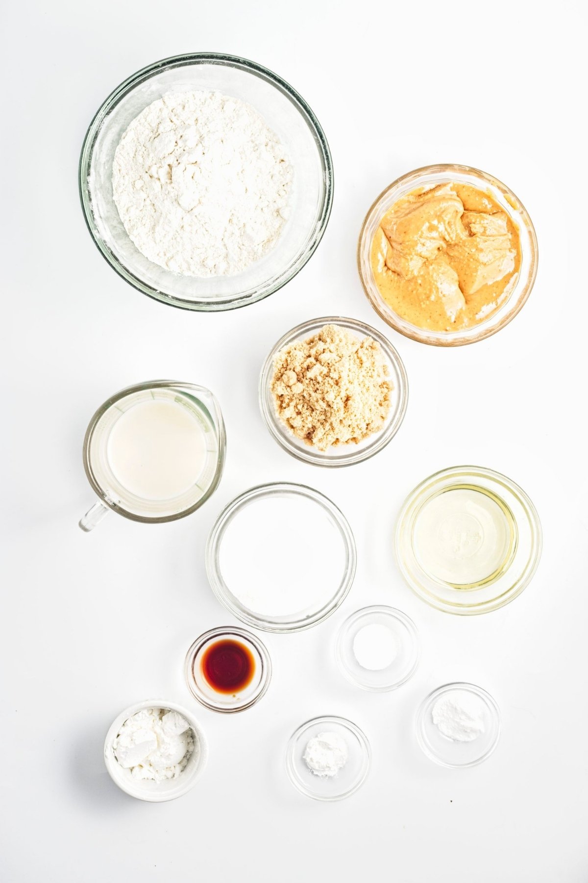 Overhead view of flour, peanut butter, brown sugar, oat milk, oil, vanilla, and small bowls of baking ingredients.