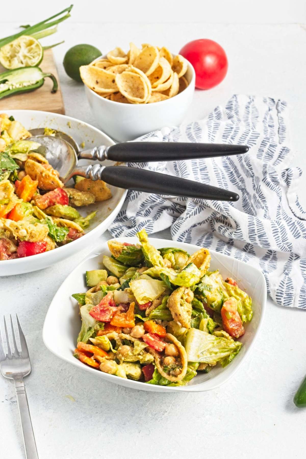 Small serving of mixed salad in a white bowl beside salad tongs and tortilla chips.