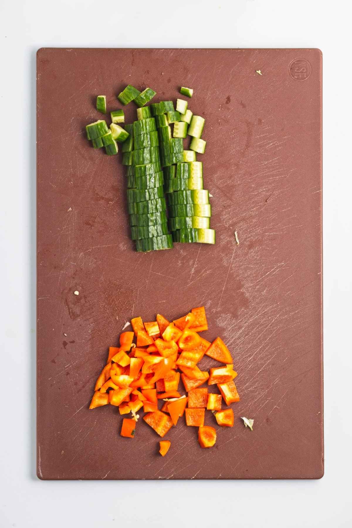 Cubed cucumber and diced orange bell pepper on a brown cutting board.