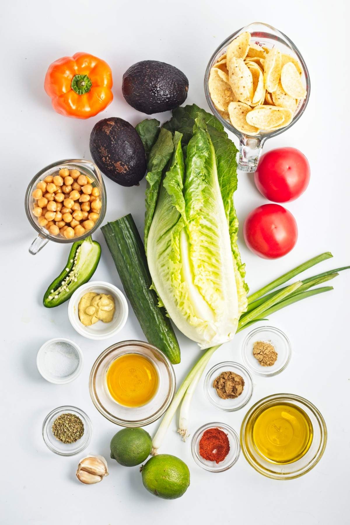 Fresh vegetables, chickpeas, tortilla chips, spices, and condiments arranged on a white surface.