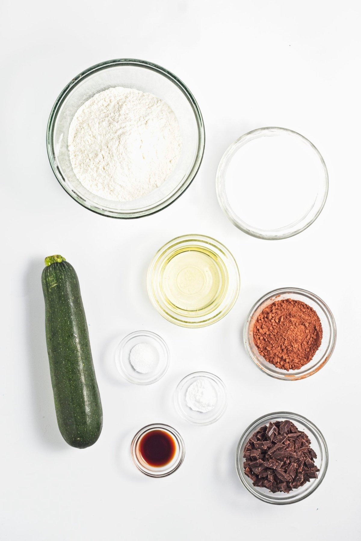 Glass bowls with flour, sugar, cocoa powder, chopped chocolate, oil, salt, baking soda, vanilla, and a whole zucchini.