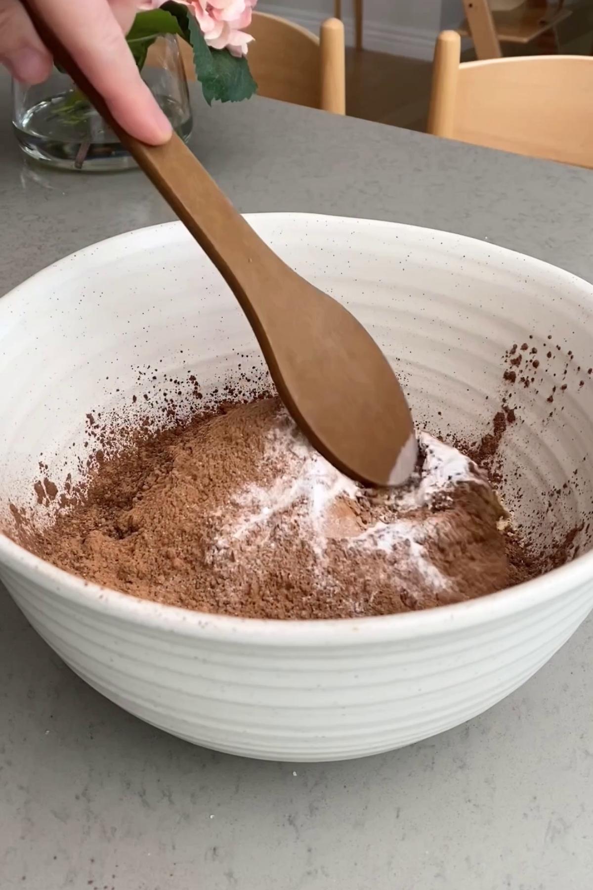 Wooden spoon mixing cocoa powder, oats, and baking ingredients in a white bowl.