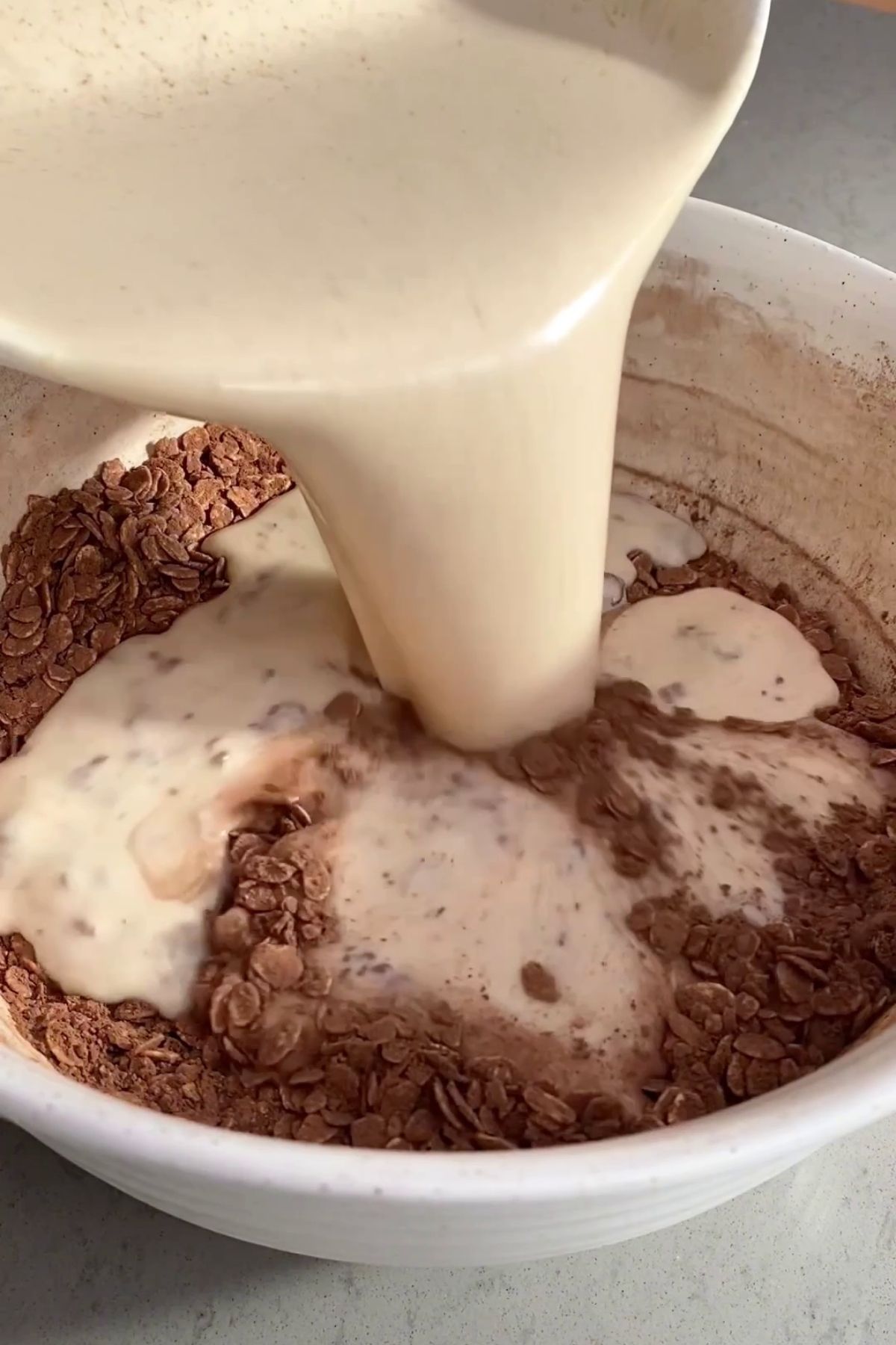 Plant milk mixture being poured over oats and cocoa powder in a mixing bowl.