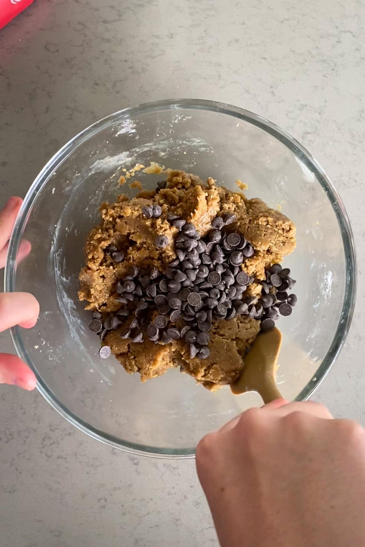 Mixing chocolate chips into soft cookie dough in a glass bowl.