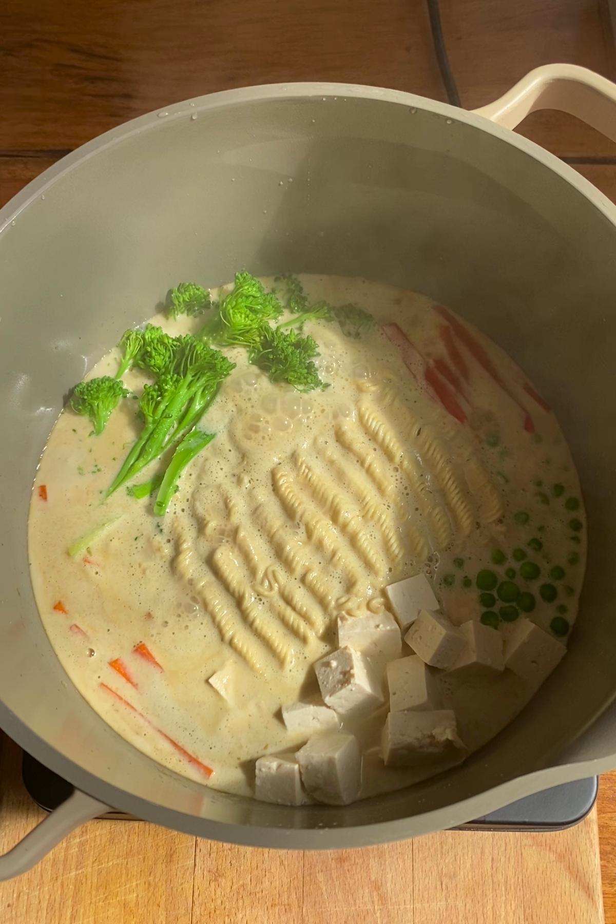 Simmering pot with noodles, broccolini, tofu cubes, peas, and peppers.