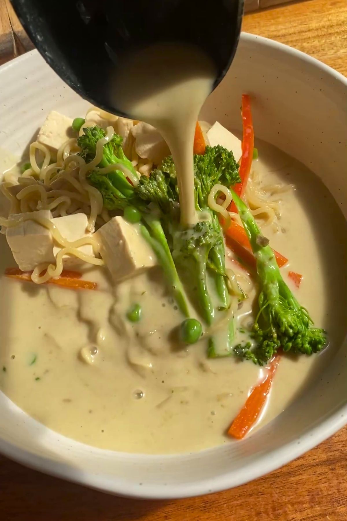 Creamy broth being ladled over noodles, tofu, and vegetables in a bowl.