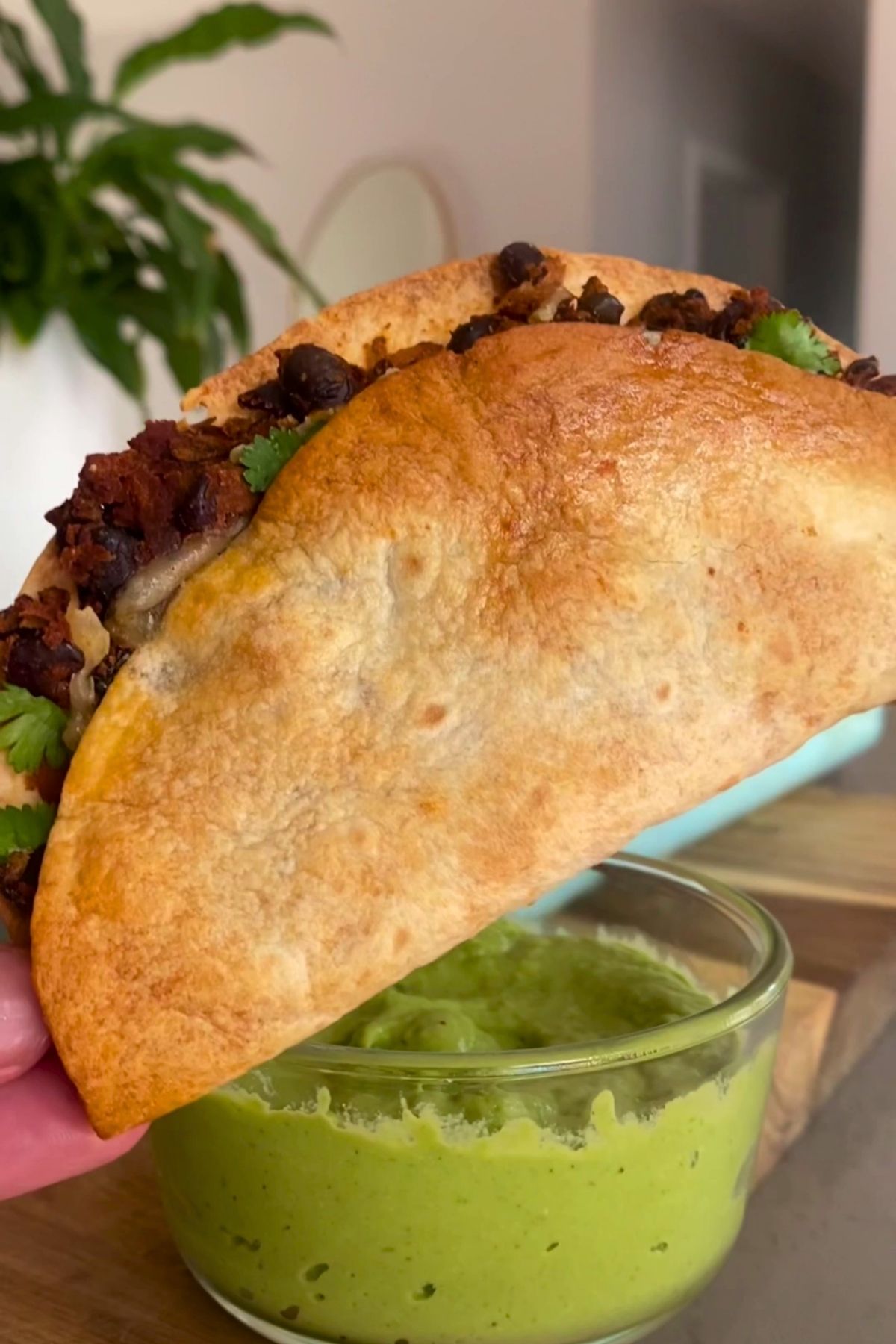 Close-up of a crispy taco with beans and herbs beside avocado dip.