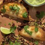 Crispy tacos topped with herbs and lime wedges beside avocado dip.