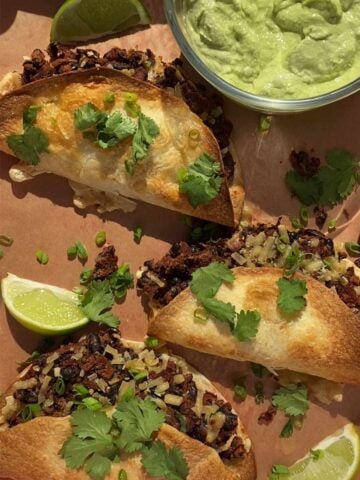 Crispy tacos topped with herbs and lime wedges beside avocado dip.