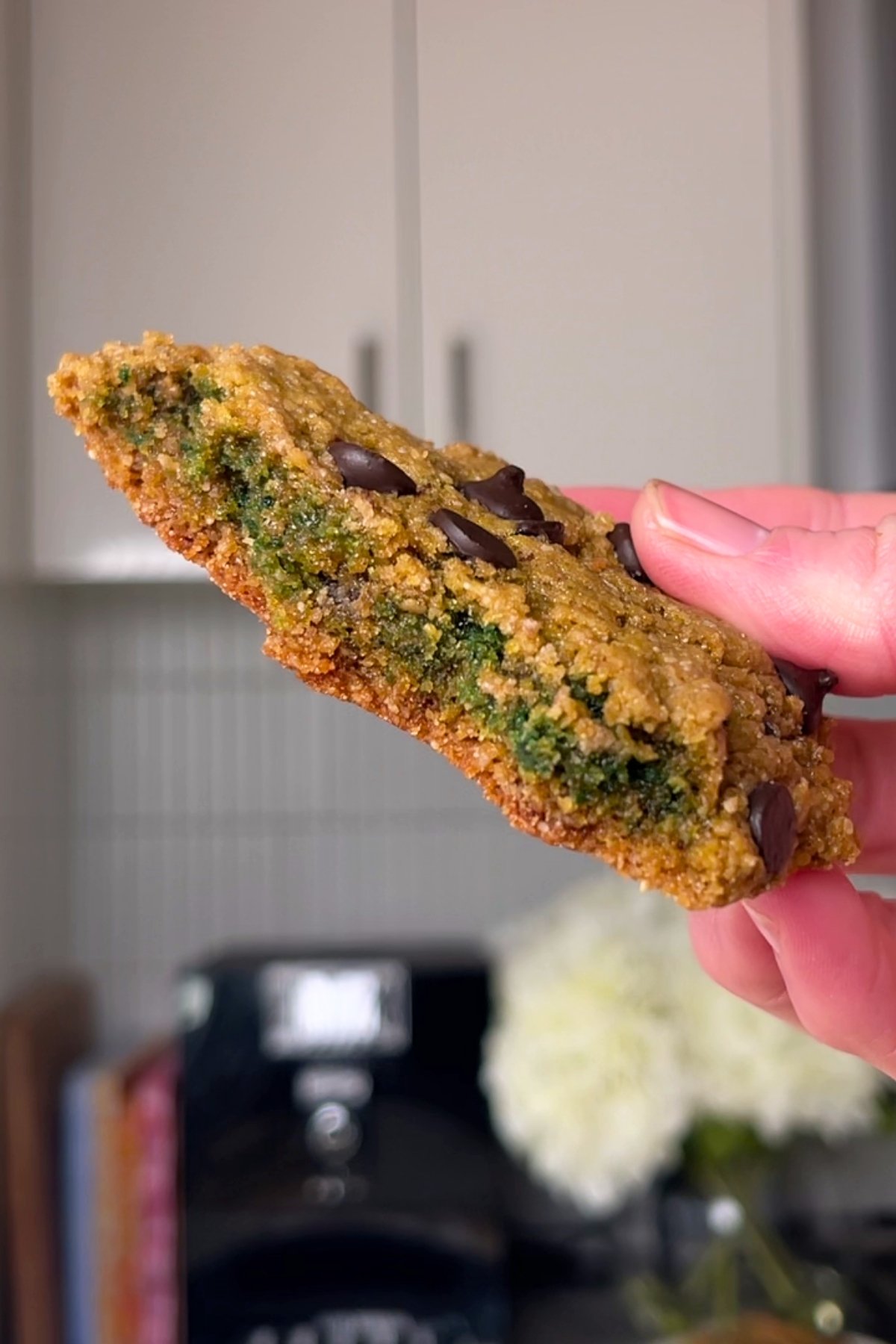 Hand holding a baked cookie with a green center and chocolate chips on top.