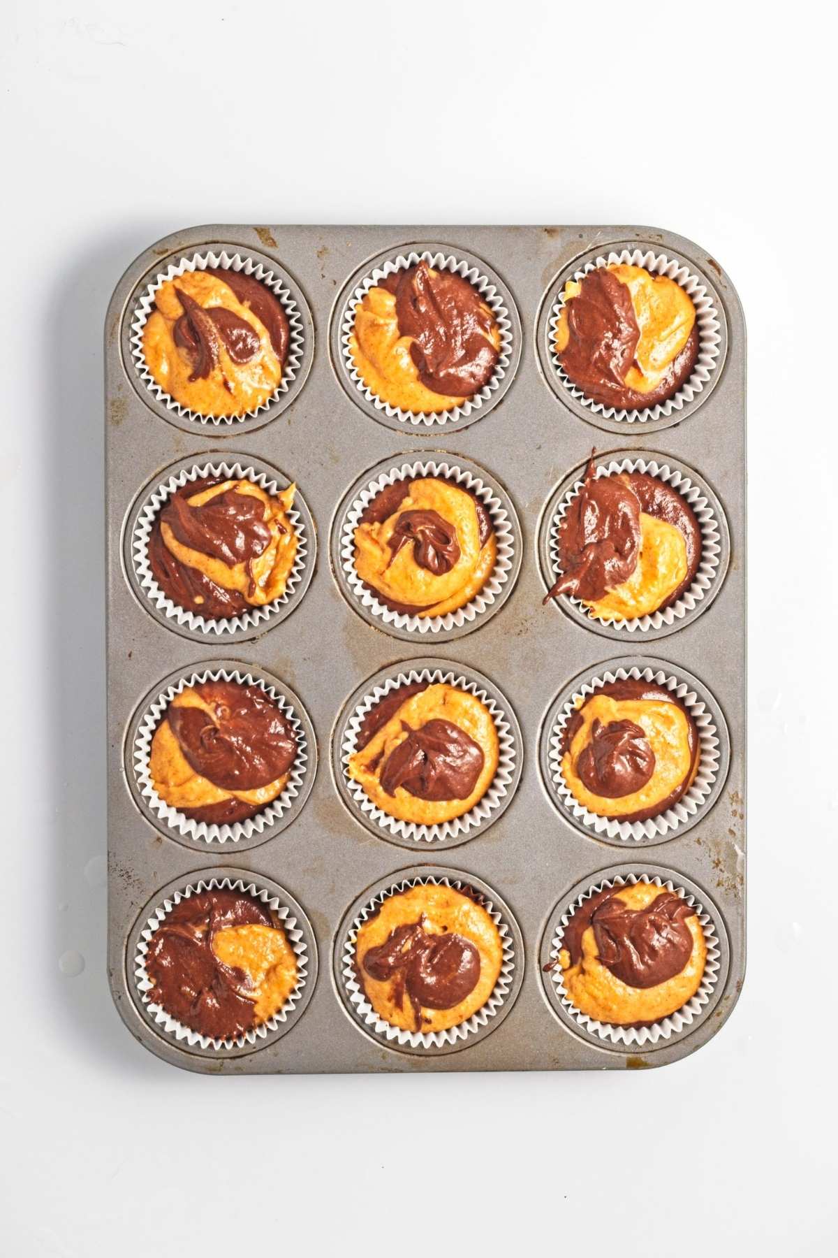 Muffin tin with liners filled with pumpkin and chocolate batter.