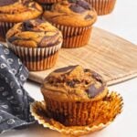 Muffin with wrapper peeled back in front of pumpkin chocolate swirl muffins on a wooden board.