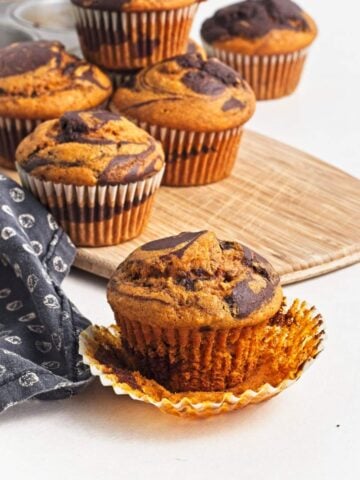 Muffin with wrapper peeled back in front of pumpkin chocolate swirl muffins on a wooden board.
