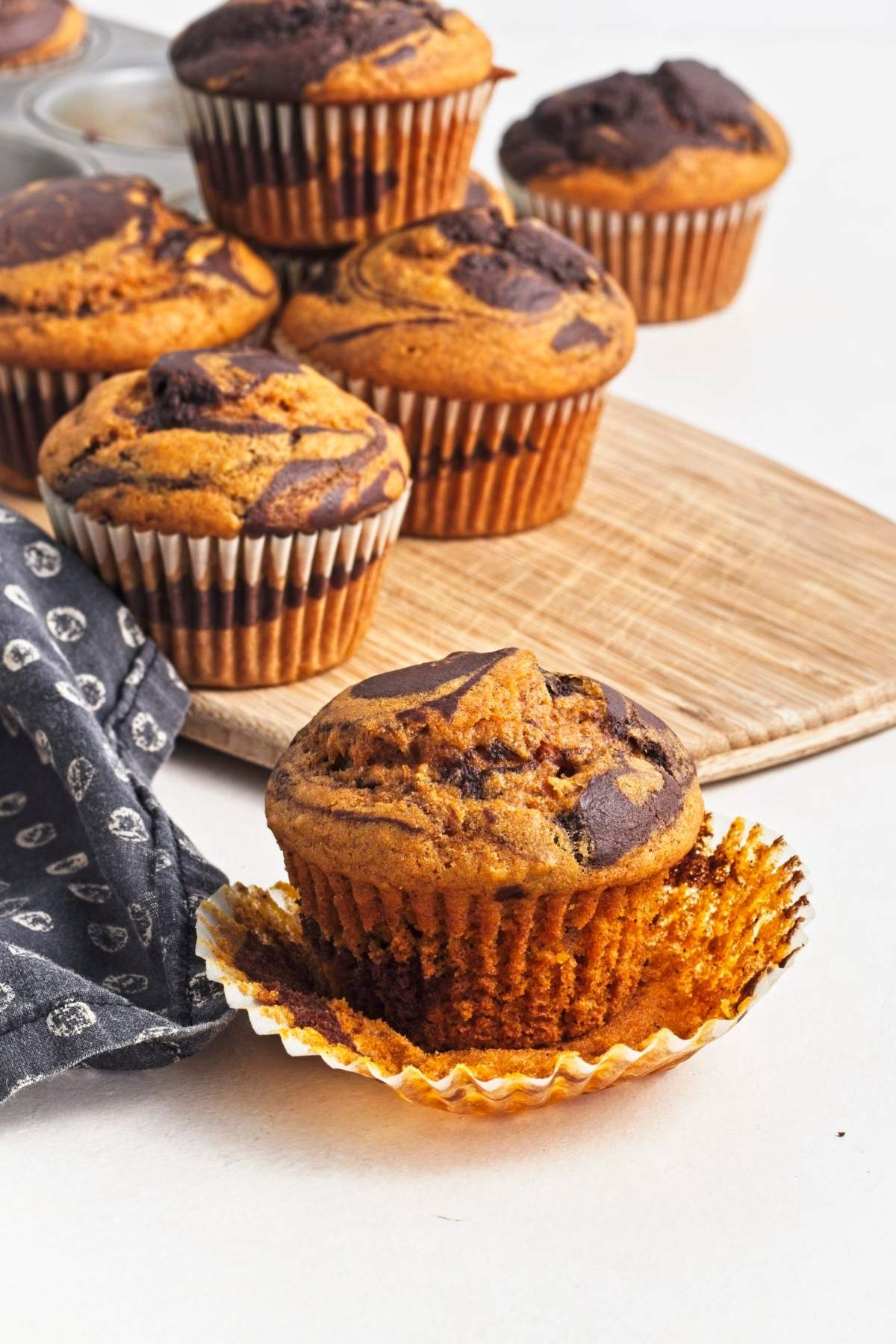 Muffin with wrapper peeled back in front of pumpkin chocolate swirl muffins on a wooden board.