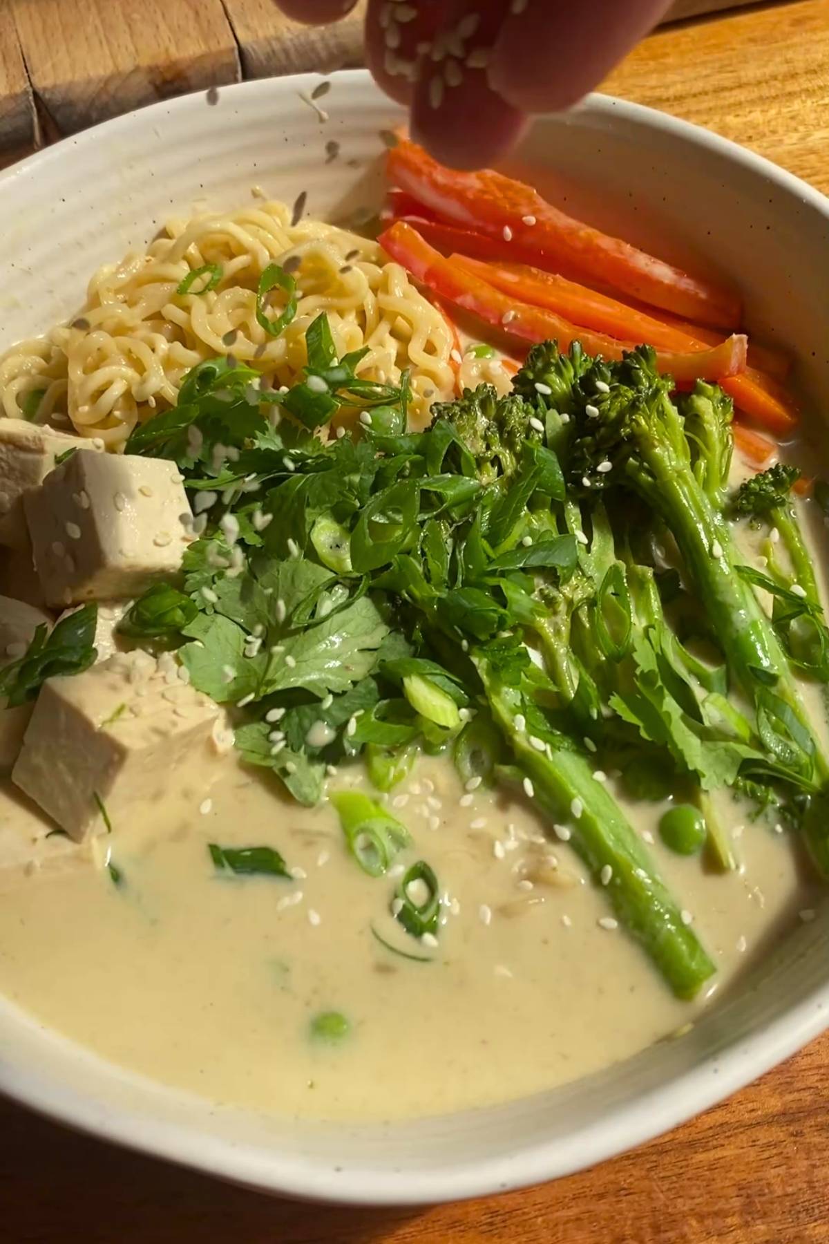 Noodle bowl topped with tofu, broccolini, peppers, herbs, and sesame seeds.
