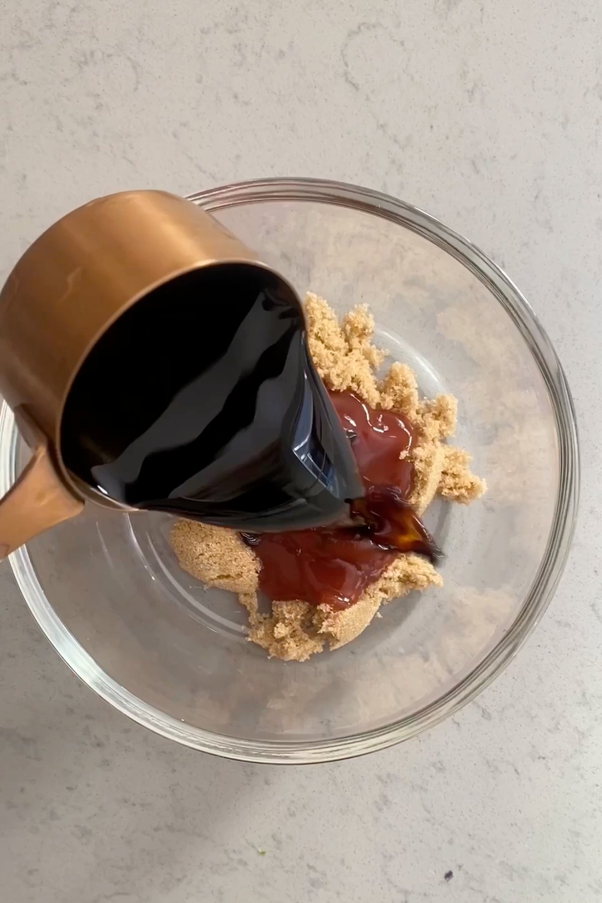 Dark soy sauce being poured from a copper measuring cup into a bowl with brown sugar and ketchup.
