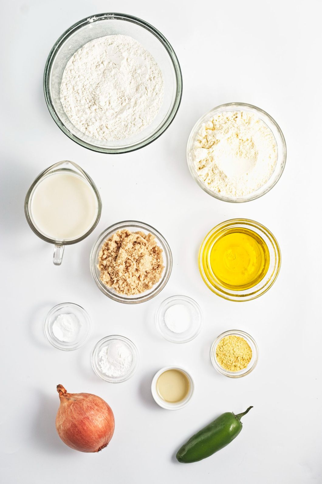 Bowls of flour, cornmeal, brown sugar, oil, oat milk, onion, jalapeño, and seasonings on a white surface.