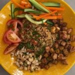 A bowl filled with fried rice, crispy tofu, pickled vegetables, tomato wedges, and chopped nuts.