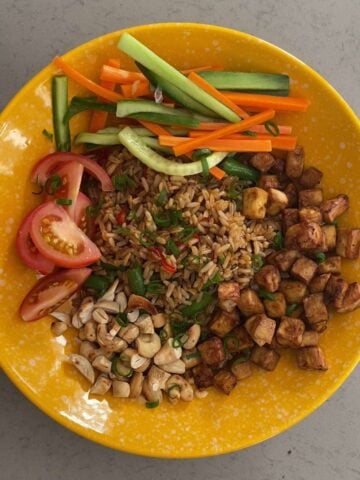 A bowl filled with fried rice, crispy tofu, pickled vegetables, tomato wedges, and chopped nuts.