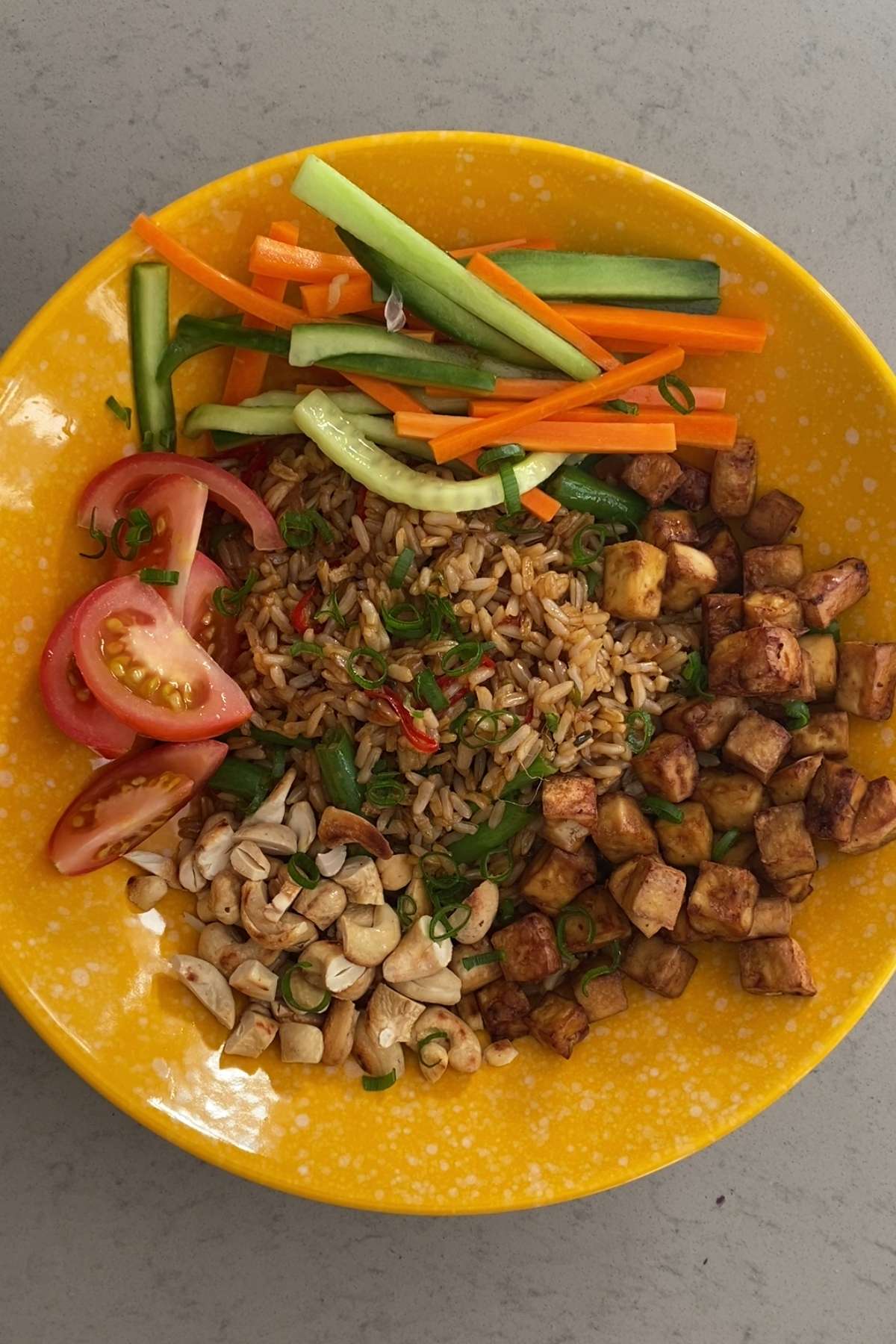 A bowl filled with fried rice, crispy tofu, pickled vegetables, tomato wedges, and chopped nuts.