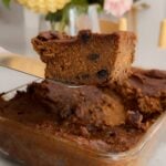 Close-up of a thick, fudgy chocolate pumpkin slice being lifted from a glass baking dish, with soft flowers blurred in the background.