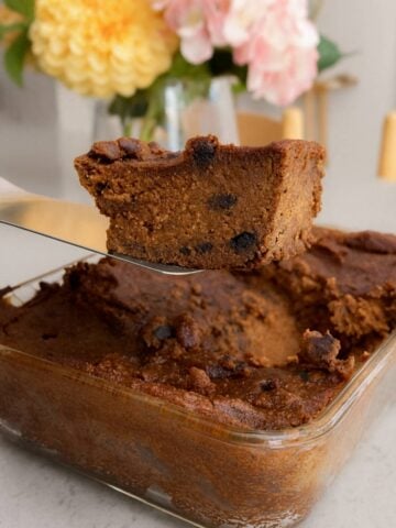 Close-up of a thick, fudgy chocolate pumpkin slice being lifted from a glass baking dish, with soft flowers blurred in the background.
