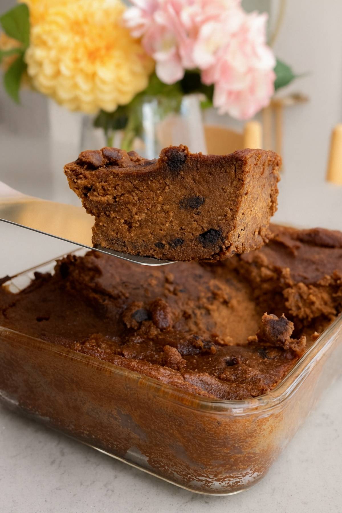 Close-up of a thick, fudgy chocolate pumpkin slice being lifted from a glass baking dish, with soft flowers blurred in the background.