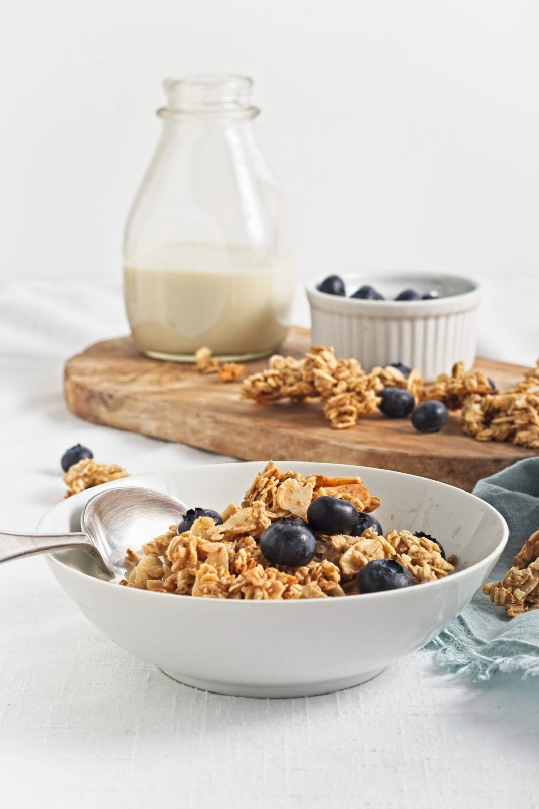 Granola with blueberries and milk served in a white bowl, with a wooden board and bottle in background.