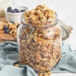 Glass jar filled with golden almond granola beside scattered clusters and a blue linen napkin.