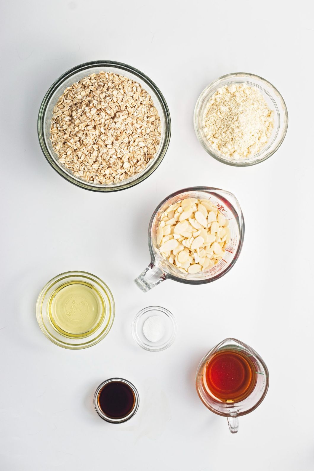 Rolled oats, almond flour, sliced almonds, maple syrup, vanilla, oil, and salt arranged in glass bowls.