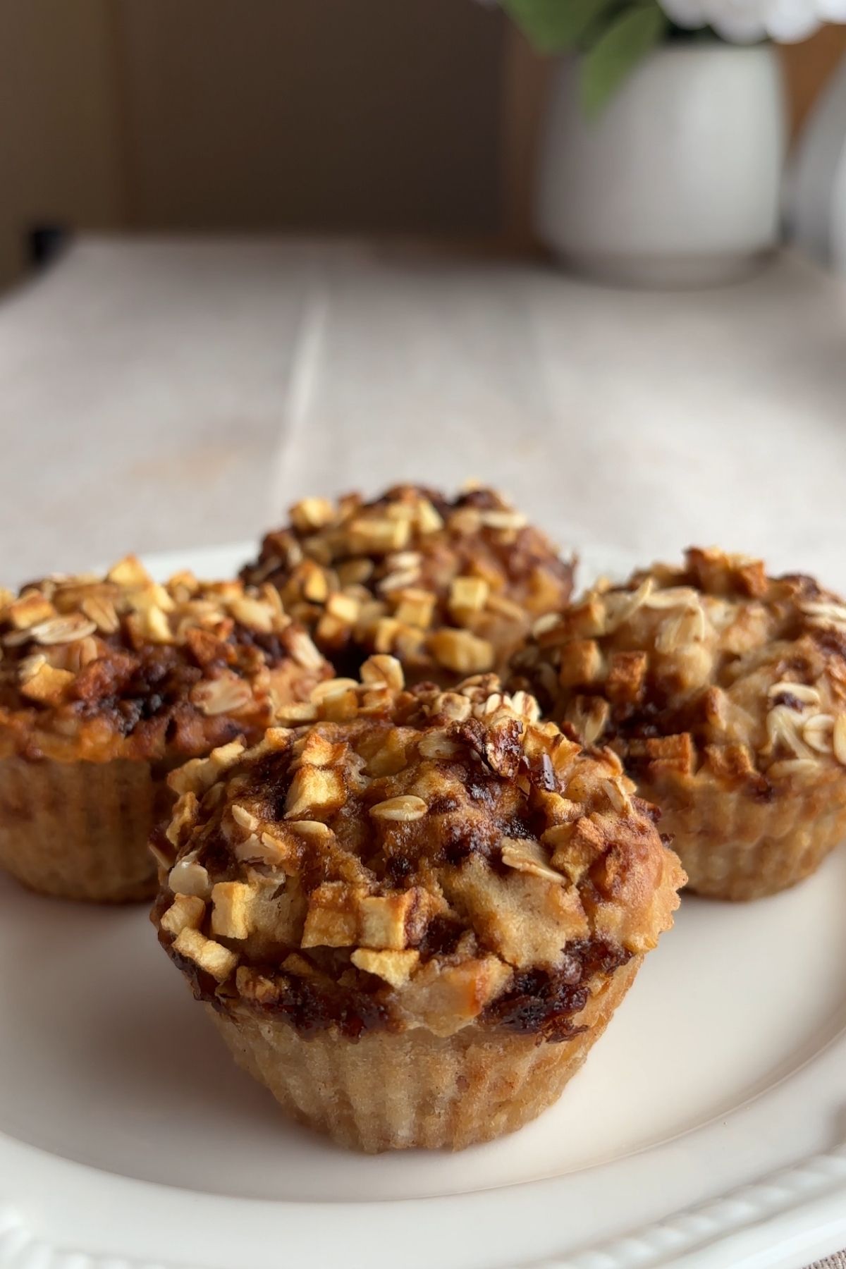 Four muffins with oats and diced apple on top arranged on a white plate.