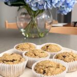 A plate of freshly baked banana muffins in paper liners on a kitchen counter with flowers in the background.