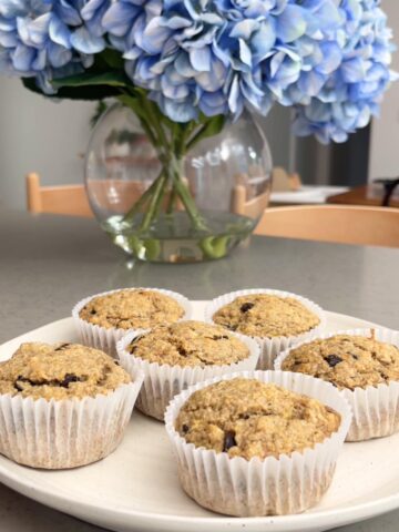 A plate of freshly baked banana muffins in paper liners on a kitchen counter with flowers in the background.