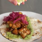 Hand squeezing a lime wedge over a taco topped with cauliflower and pink pickled onions.