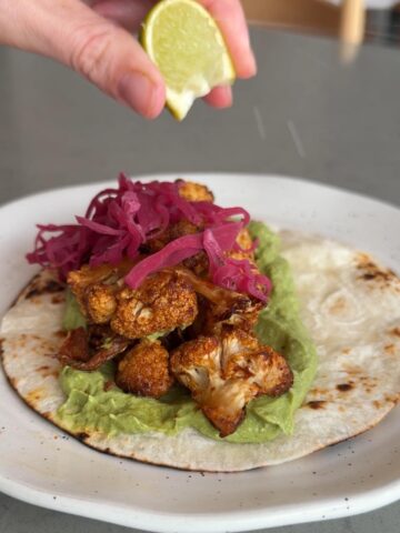 Hand squeezing a lime wedge over a taco topped with cauliflower and pink pickled onions.