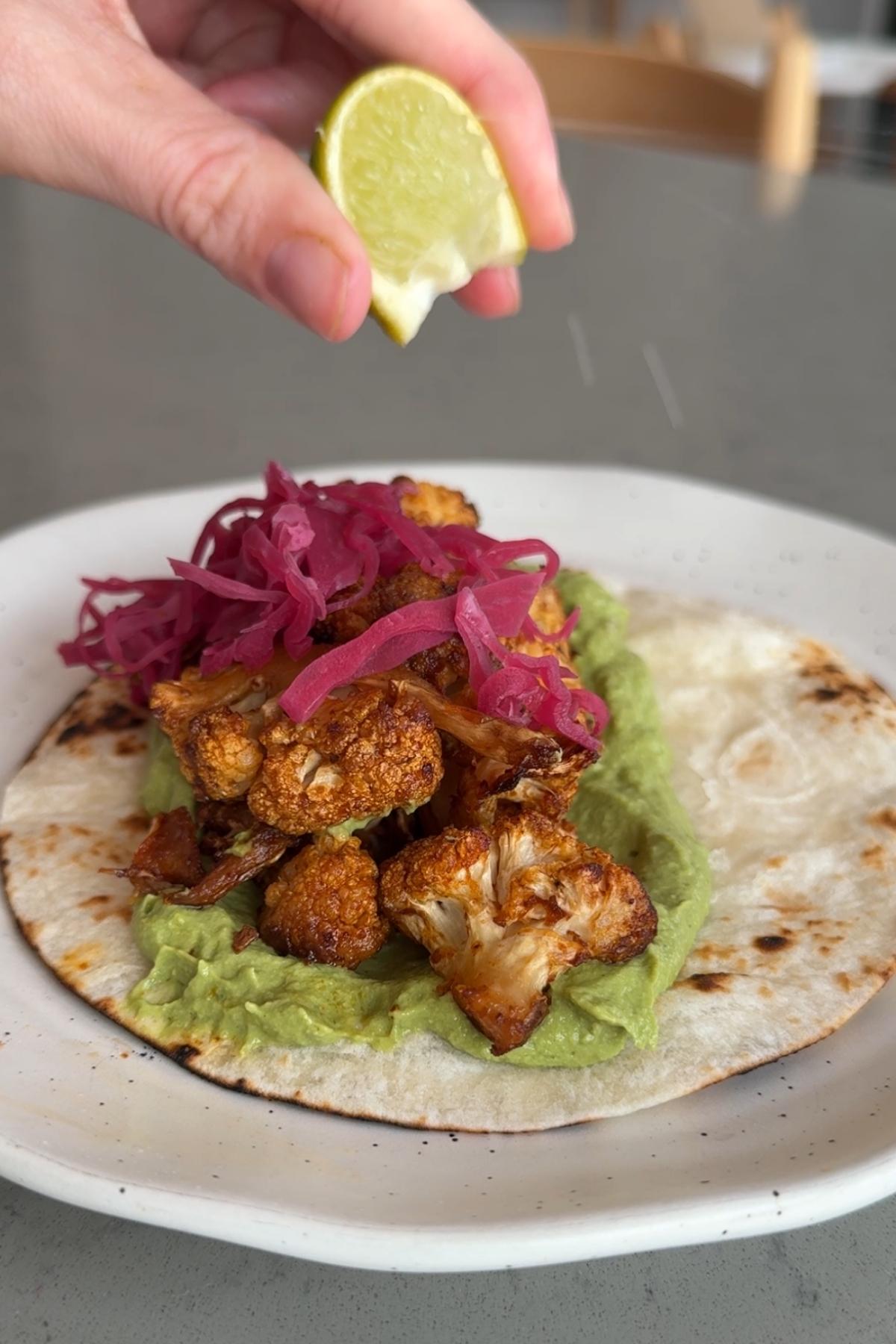 Hand squeezing a lime wedge over a taco topped with cauliflower and pink pickled onions.