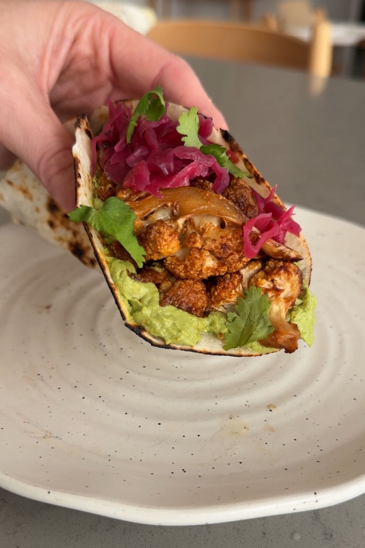 Hand holding a folded taco filled with roasted cauliflower, pink pickled onions, and herbs on a white plate.
