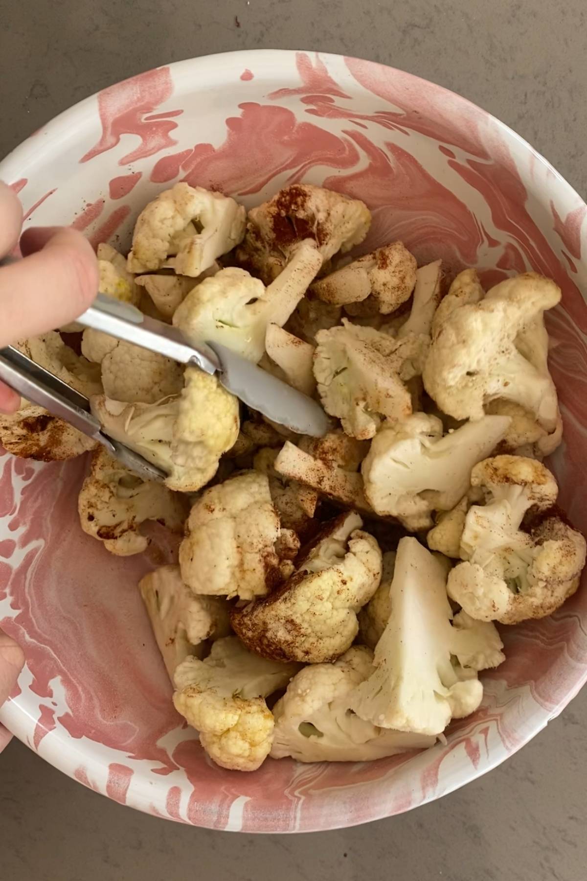 Seasoned cauliflower florets being tossed in a bowl with tongs.