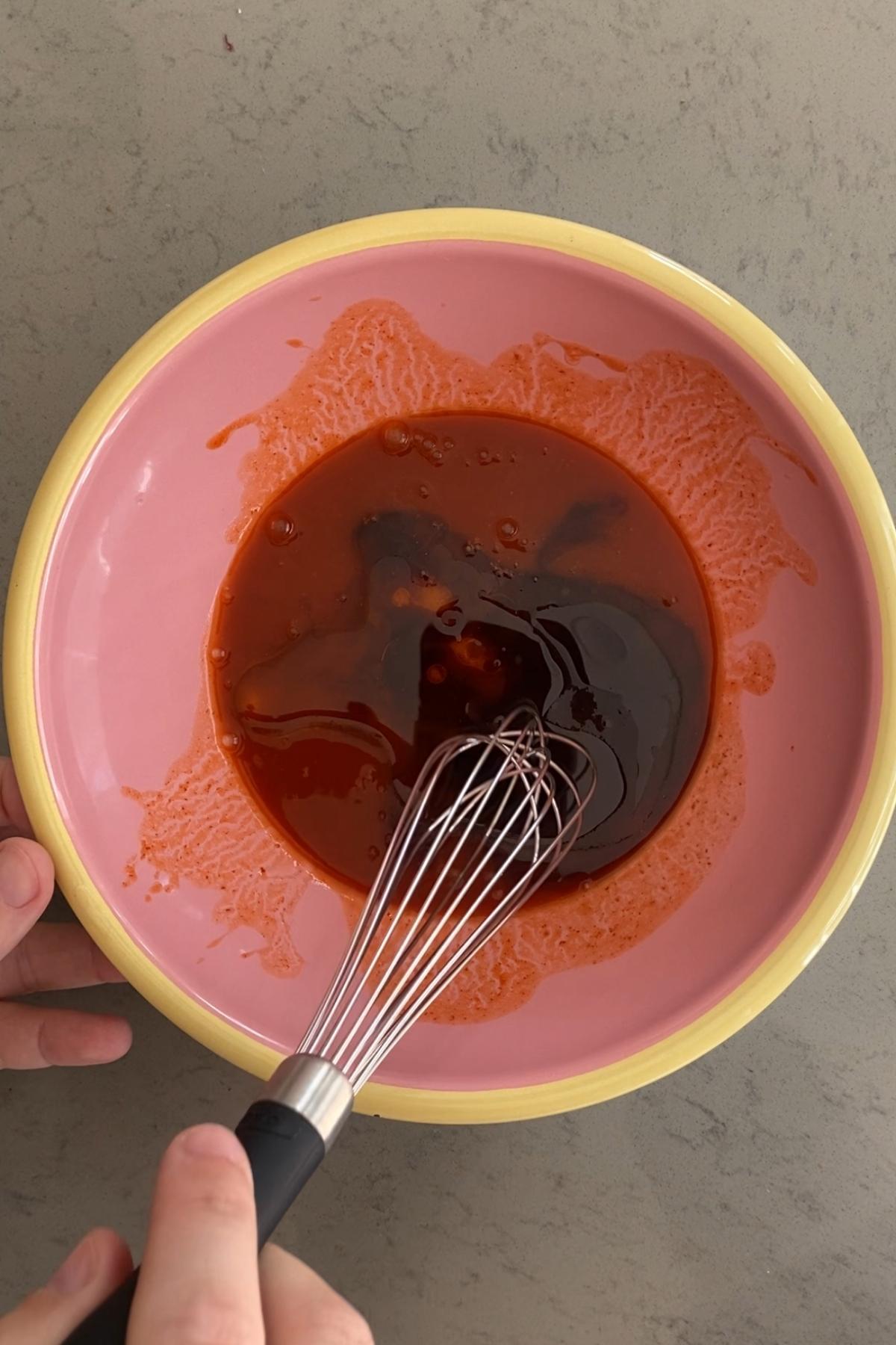 A whisk mixing a reddish sauce in a bowl.