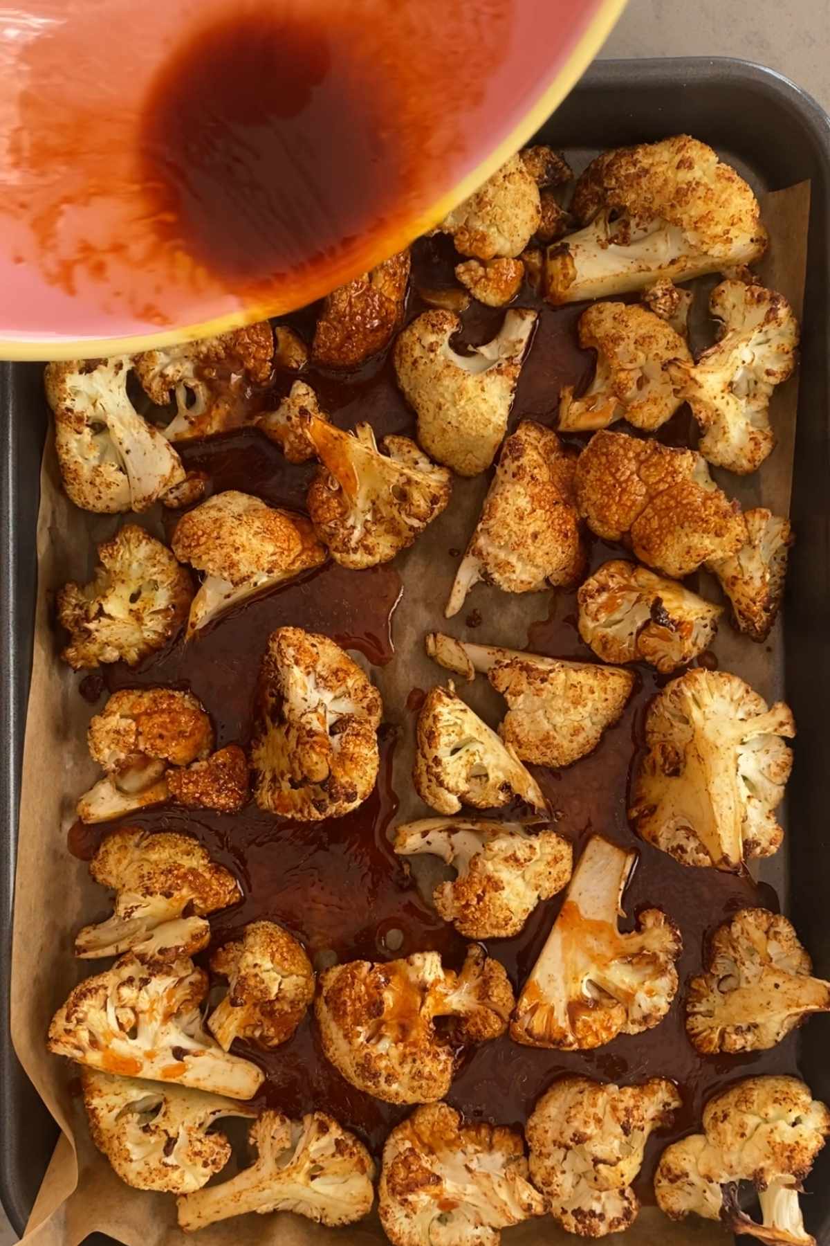 Roasted cauliflower pieces on a baking tray being coated with sauce from a bowl.