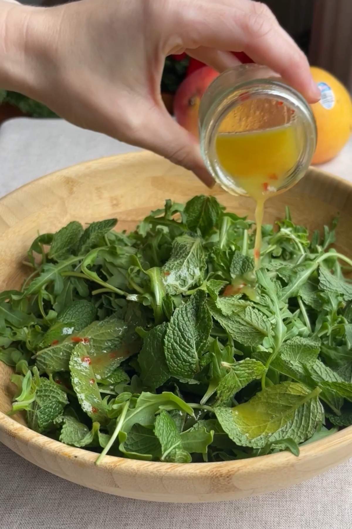 Hand pouring lime chili dressing over fresh arugula and mint leaves in a wooden bowl.