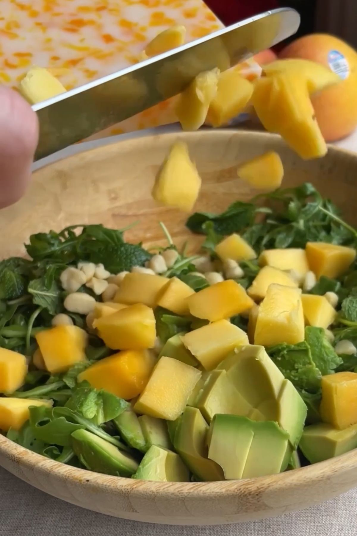 Chopped mango and avocado being added to a salad bowl with greens and macadamias.