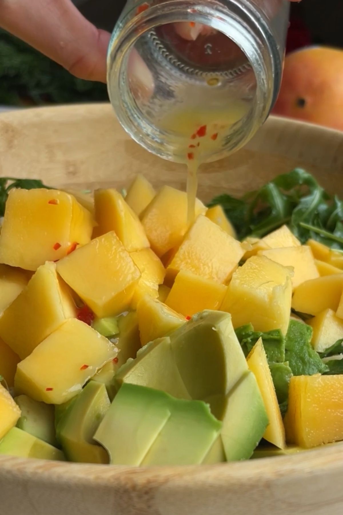 Hand pouring lime chili dressing over diced mango and avocado in a wooden salad bowl.